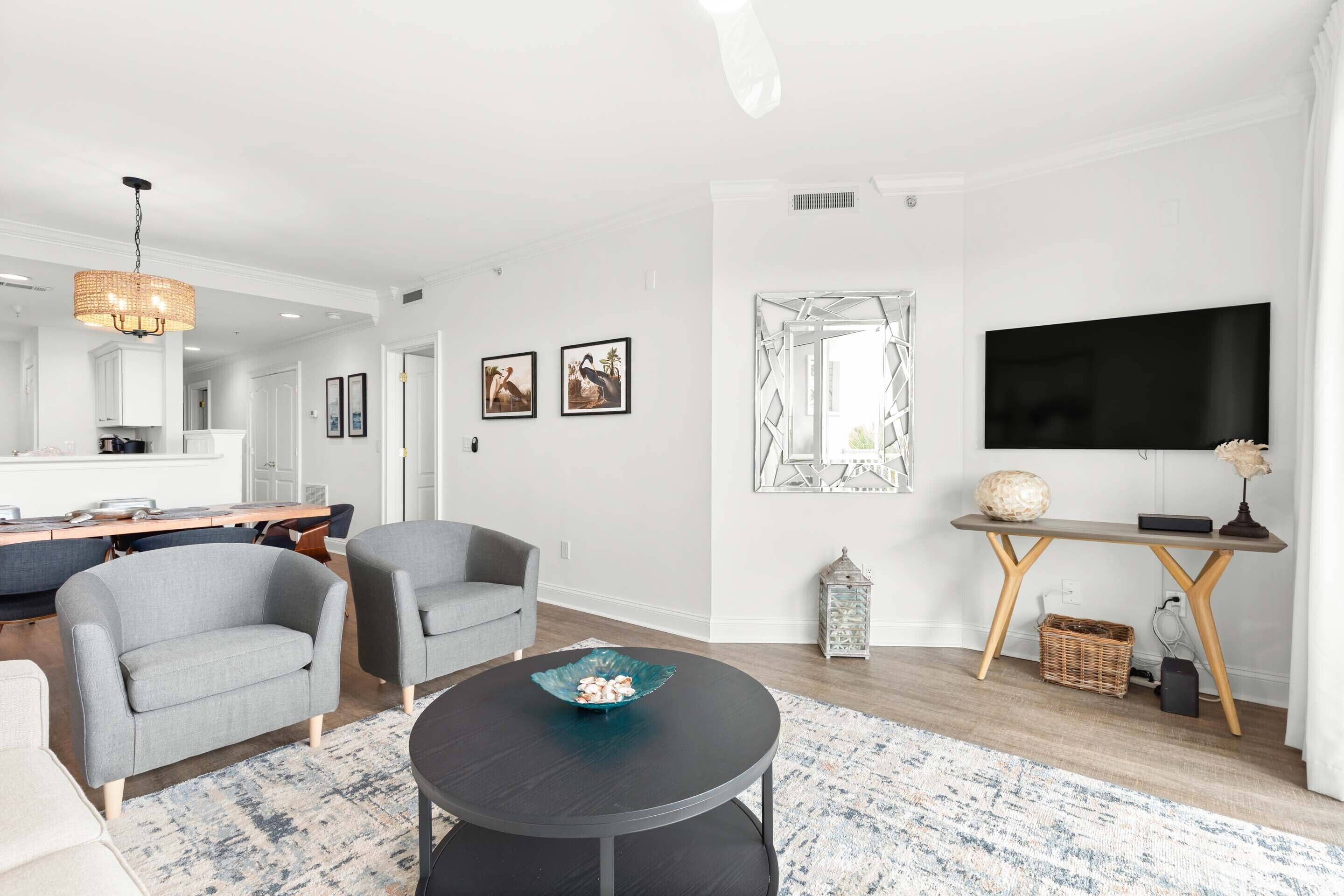 Un salon moderne avec une table basse ronde noire, des fauteuils gris, un téléviseur et un coin repas comprenant un buffet blanc et une table en bois.