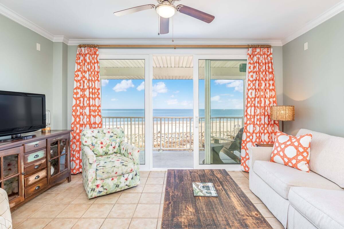 A living room with a television, a floral armchair, a sofa with orange pillows, a wooden coffee table, and a view of the ocean through large glass doors.