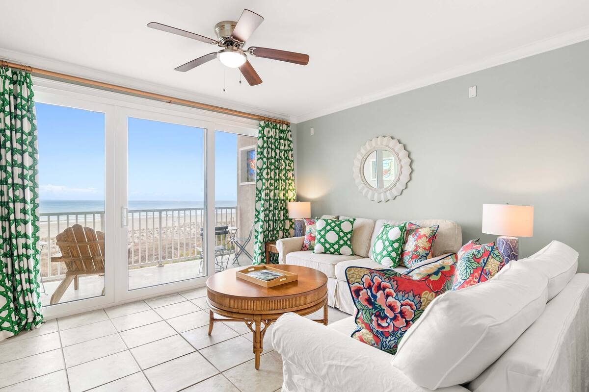 A spacious living room with a balcony, a ceiling fan, a round wooden coffee table, and a white sofa with colorful cushions.