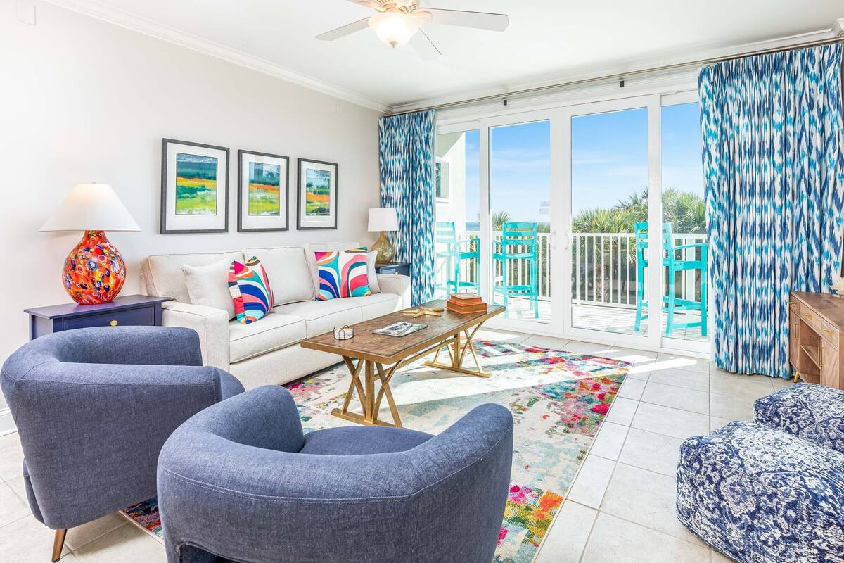A bright living room with a sofa, two armchairs, a coffee table, and a floor lamp. There are framed pictures on the wall, a colorful rug, and a view of the outside through large sliding doors.