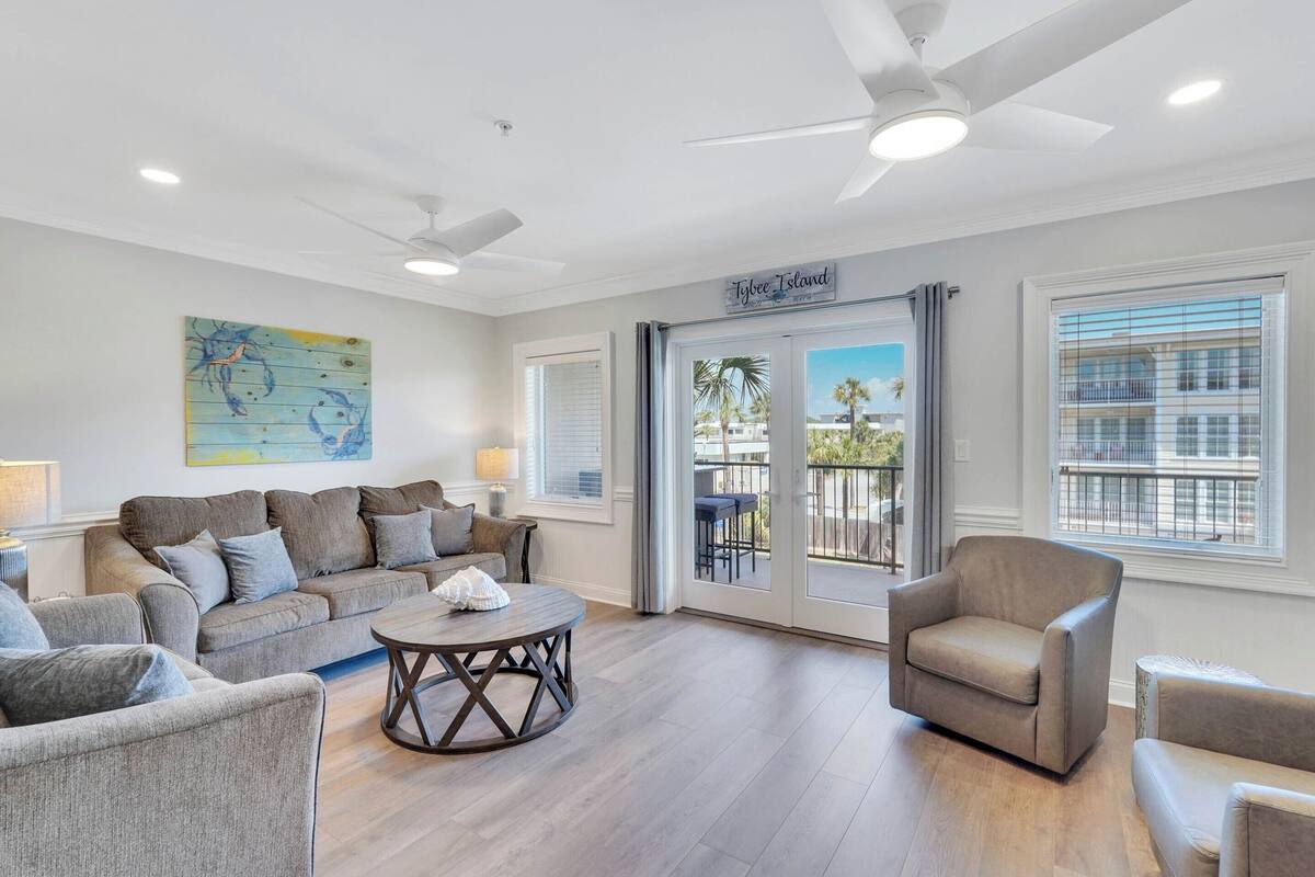 A spacious living room with a grey sofa, a round coffee table, and a beige armchair. There is a ceiling fan, a painting on the wall, and a sign that reads "Tybee Island".