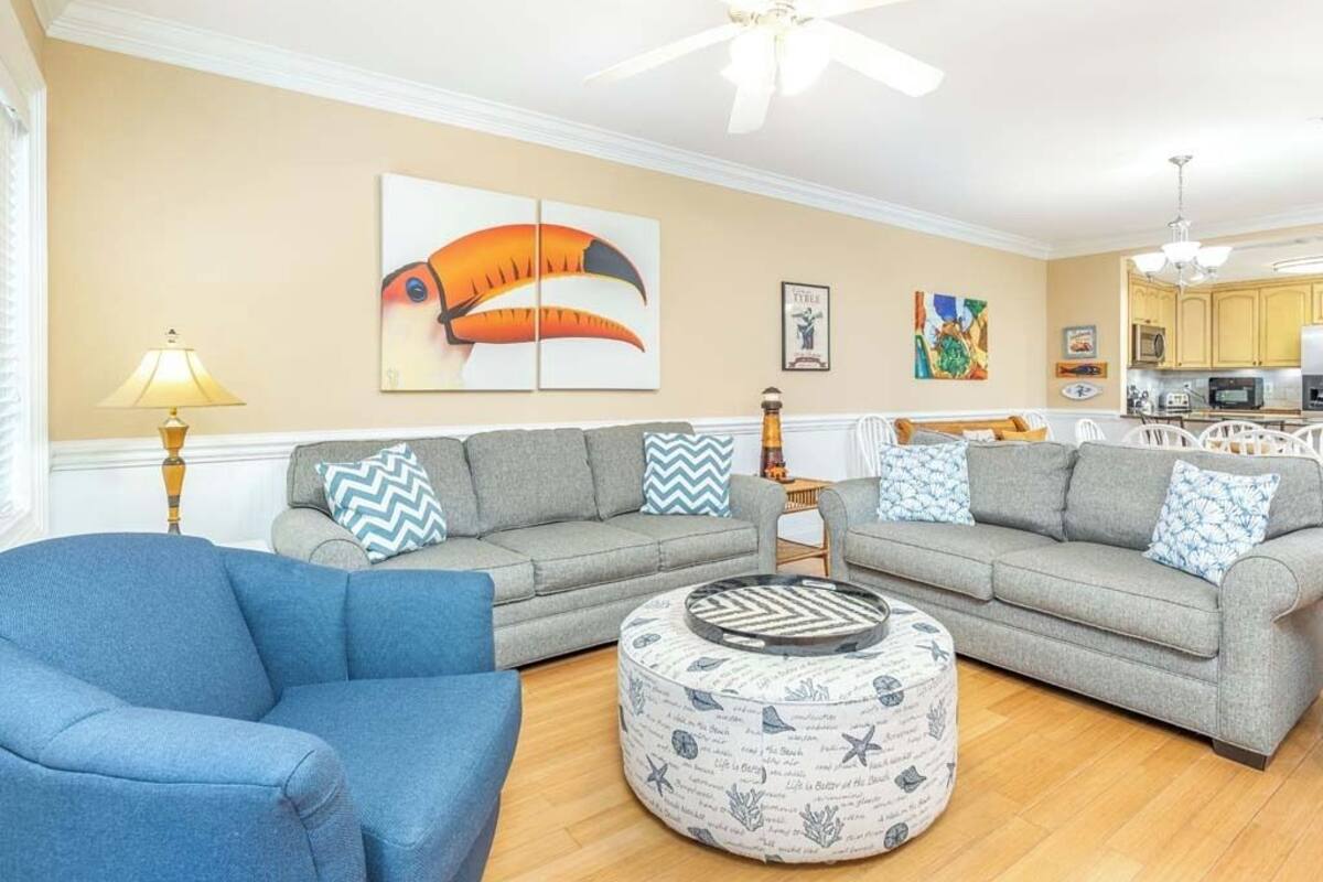 A living room with two grey sofas, a blue armchair, a round ottoman with nautical prints, and a ceiling fan.