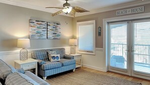 A living room with a striped sofa, a matching ottoman, a ceiling fan, and a large window with blinds. - Brass Rail Villas by Tybee Vacation Rentals (Tybee Island)