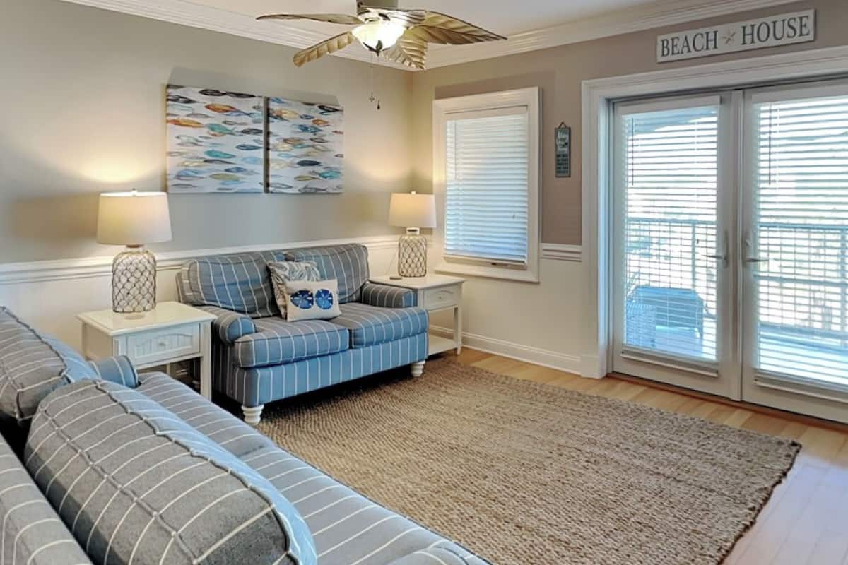 A living room with a striped sofa, a matching ottoman, a ceiling fan, and a large window with blinds.