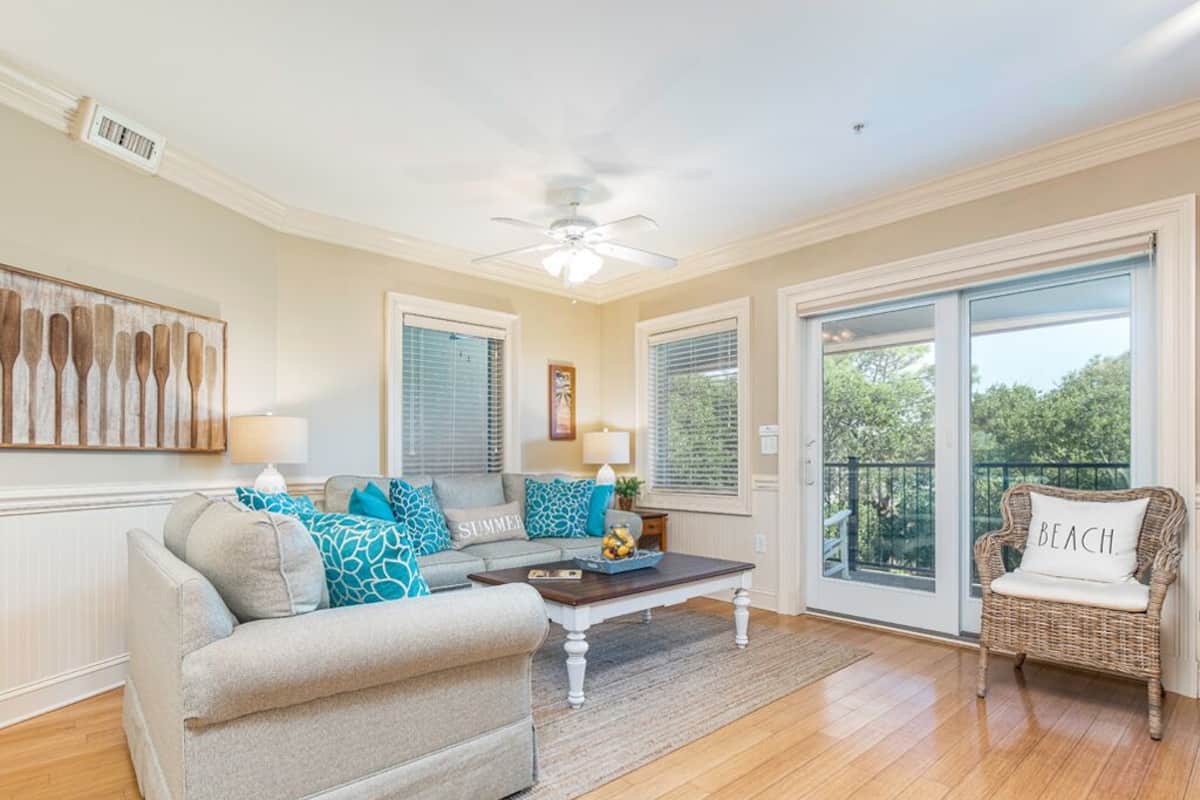A spacious living room with a grey sofa, a wicker chair, and a coffee table. There are two windows with blinds, a ceiling fan, and a large sliding glass door leading to a balcony.