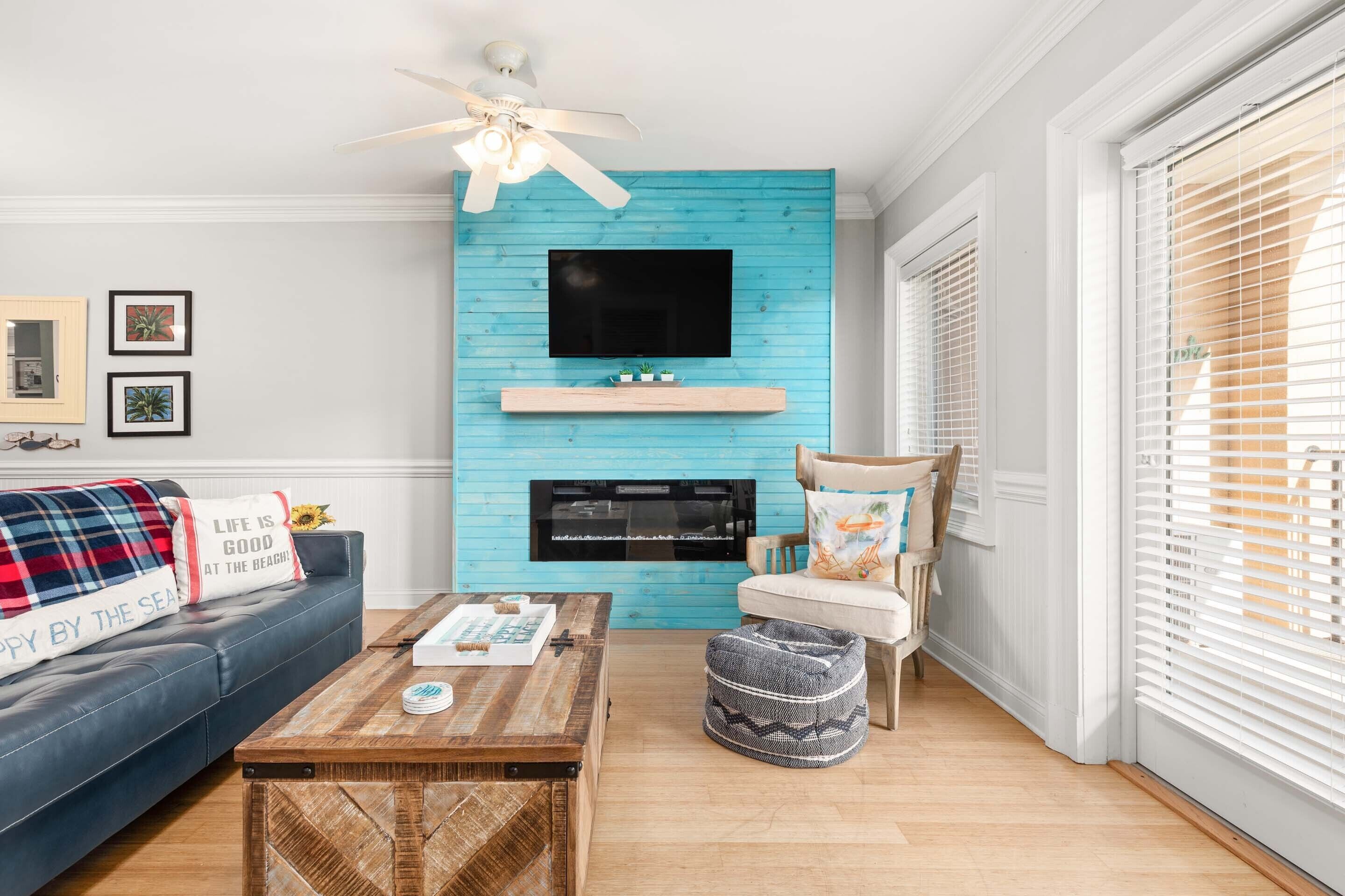 A living room with a blue accent wall, a fireplace, a television mounted above it, a sofa with a decorative pillow, a coffee table, and a chair with a cushion.