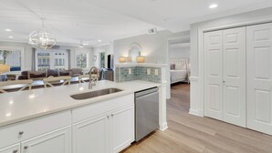 A modern kitchen with white cabinetry, a central island with a sink, and a dishwasher. There is a dining area with a table and chairs, and a bedroom with a large bed and a closet. - Brass Rail Villas by Tybee Vacation Rentals (Tybee Island)