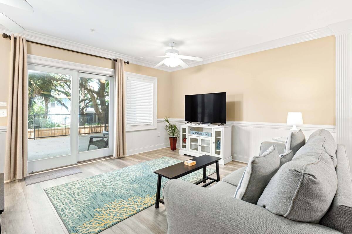 A living room with a sofa, a coffee table, a TV on a cabinet, and a sliding glass door leading to an outdoor area.