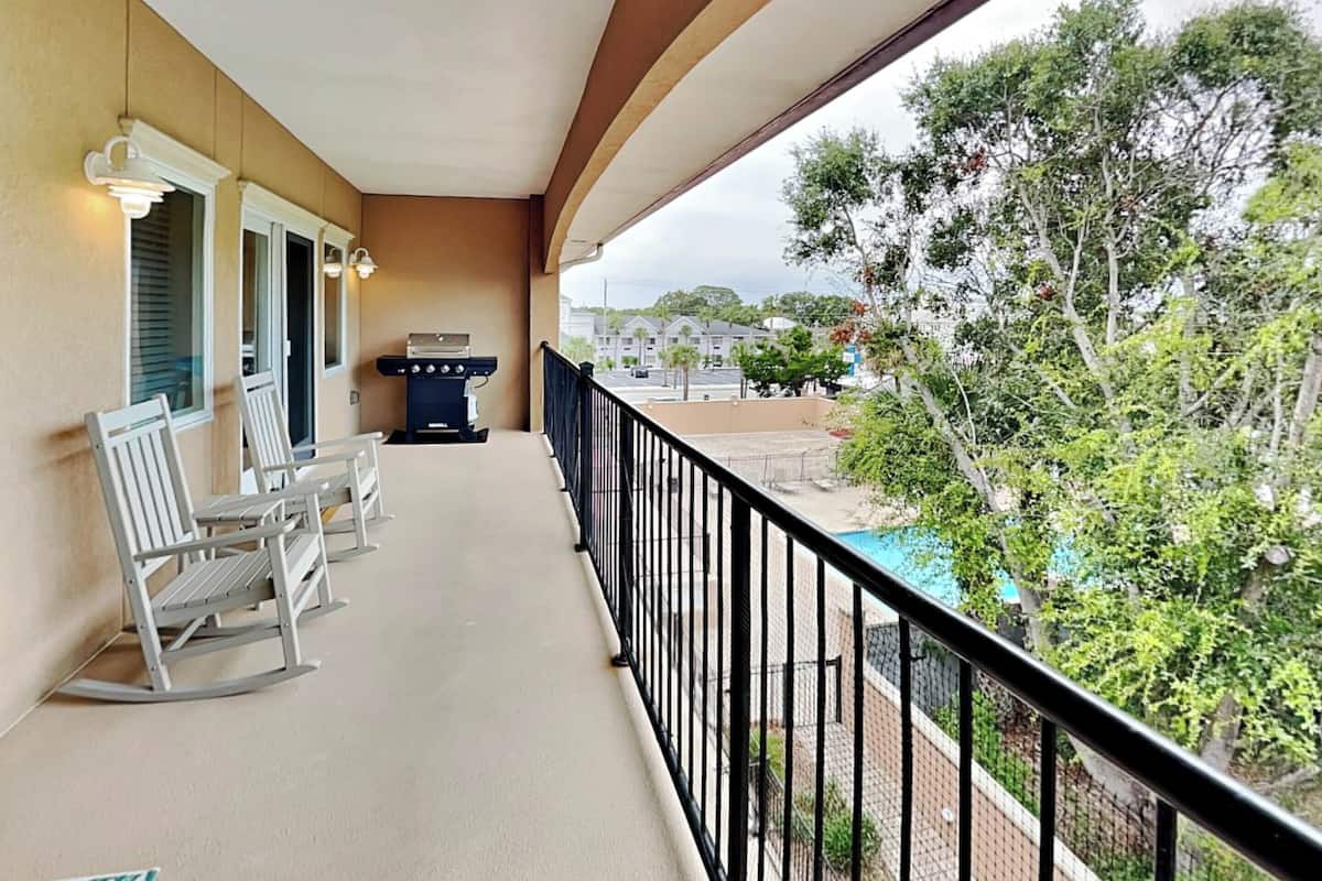 A balcony with white rocking chairs, a barbecue grill, and a view of a swimming pool and greenery.