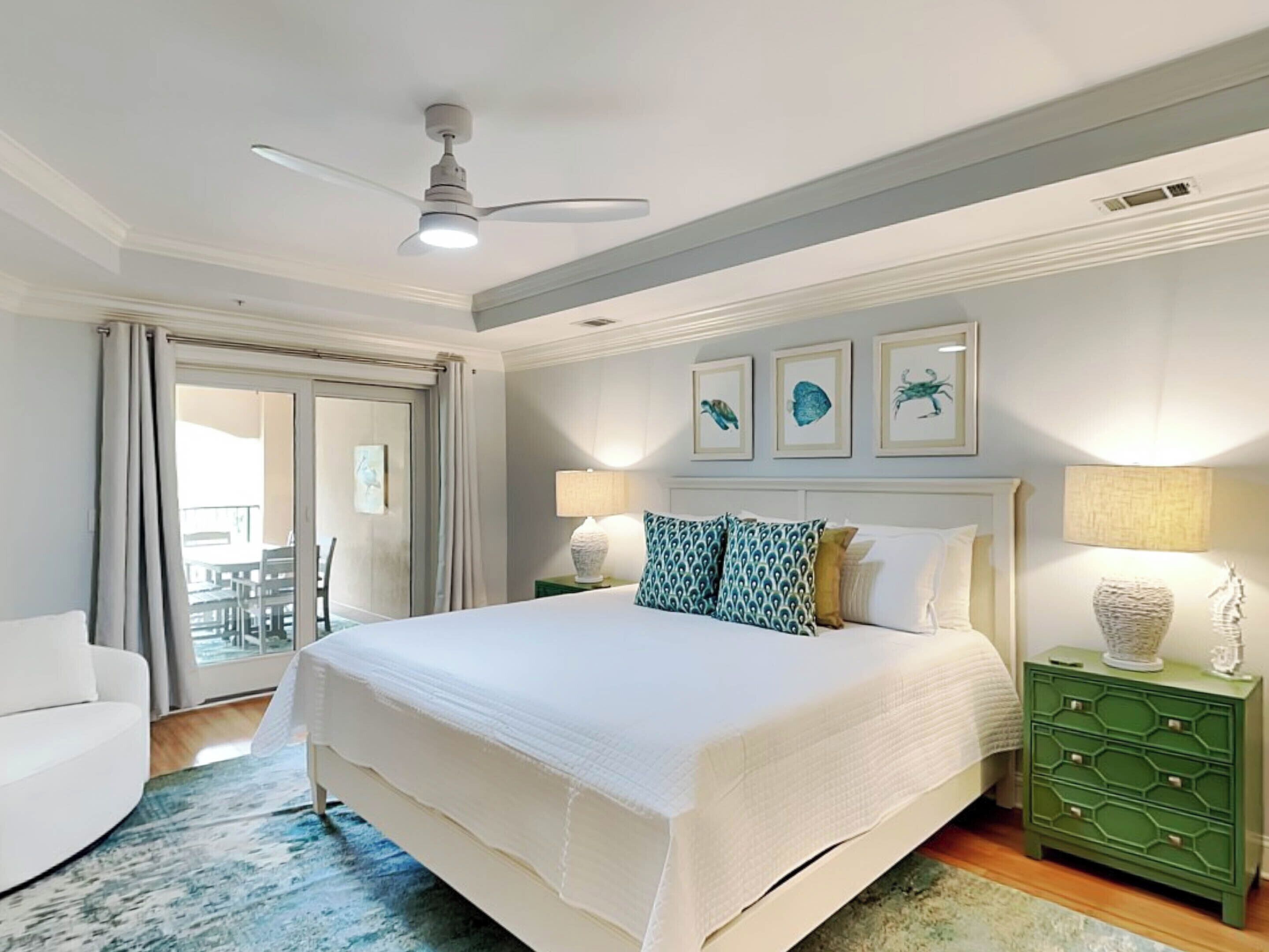 Une chambre d’hôtel avec un grand lit, un ventilateur de plafond, une table de chevet verte et une vue sur l’extérieur grâce à une porte coulissante.