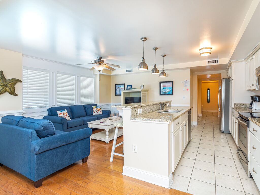 A spacious living room with a blue sofa set, a small island with a sink, and a kitchen area with white cabinets and a refrigerator.