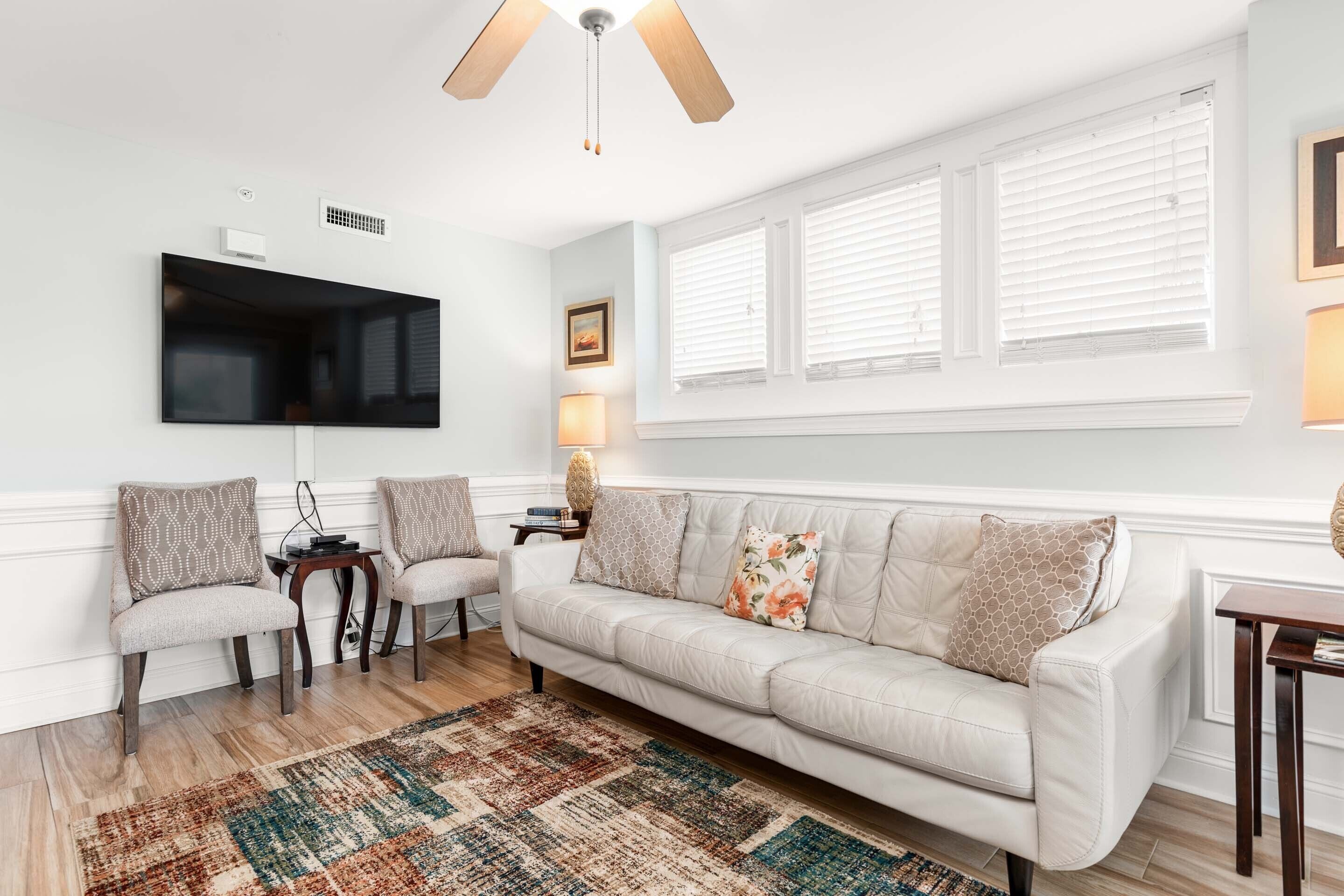 A living room with a white sofa, two armchairs, a coffee table, a flat-screen TV mounted on the wall, and a ceiling fan.