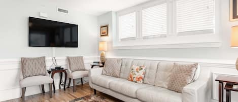A living room with a white sofa, two armchairs, a coffee table, a flat-screen TV mounted on the wall, and a ceiling fan.