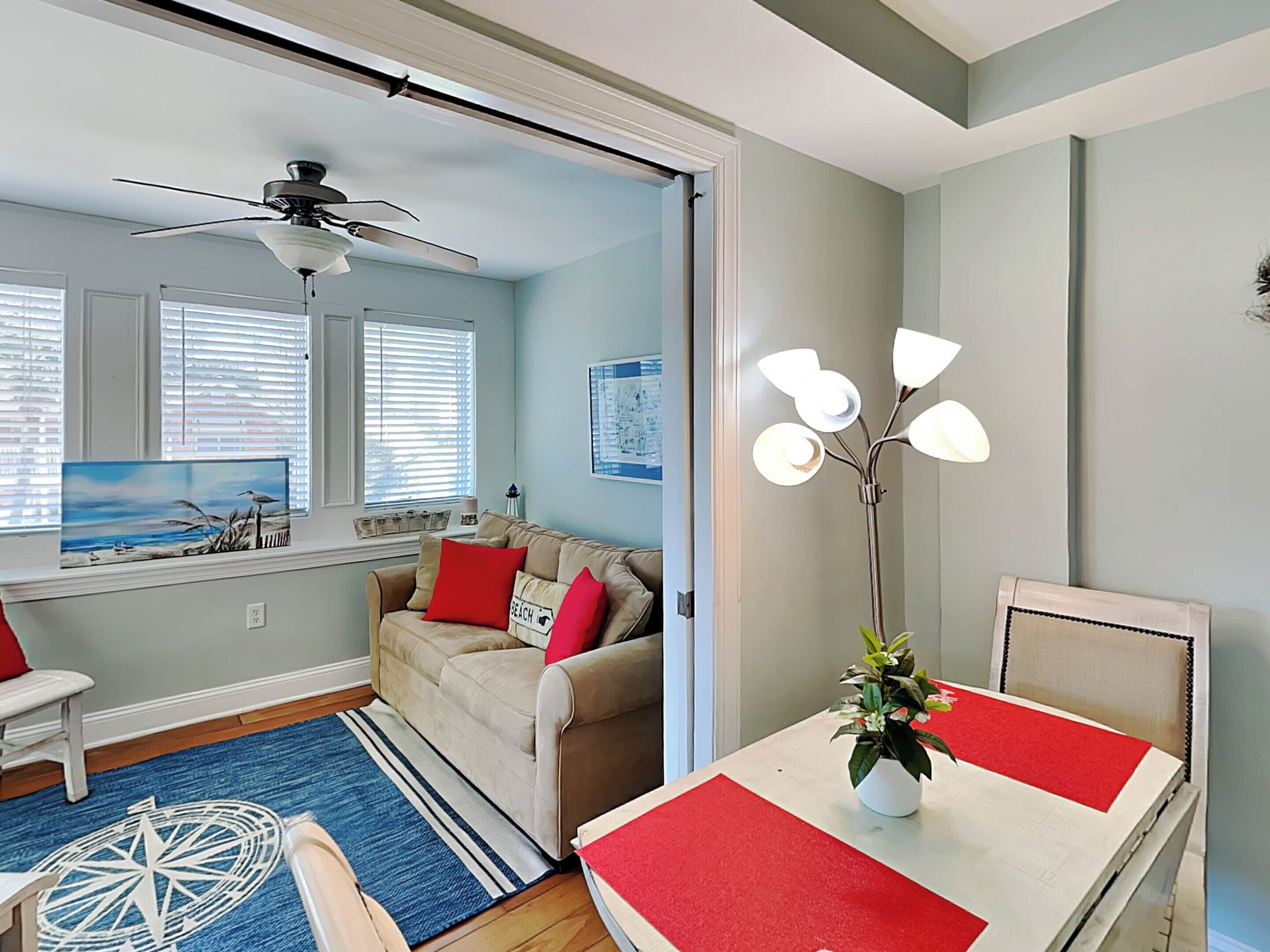 Una sala moderna con un sofá beige, cojines rojos, una mesa de comedor con individuales rojos, una lámpara de pie y un cuadro de una playa.