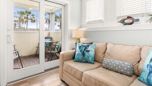 A beige leather sofa with patterned cushions, a glass door leading to a patio with outdoor furniture, and a lamp on a side table. - Captains Watch 1 by Tybee Vacation Rentals (Tybee Island)