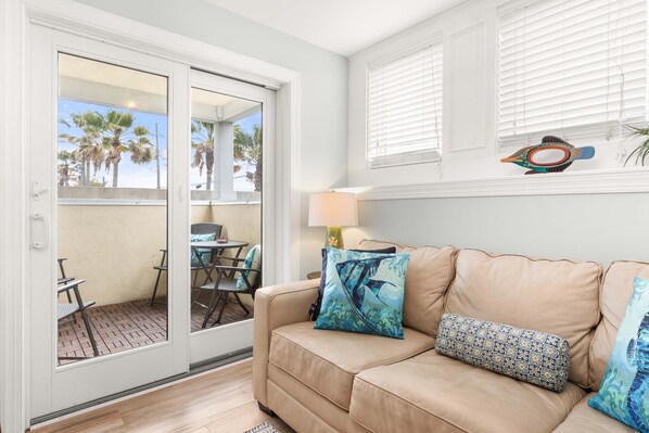 A beige leather sofa with patterned cushions, a glass door leading to a patio with outdoor furniture, and a lamp on a side table. - Captains Watch 1 by Tybee Vacation Rentals (Tybee Island)