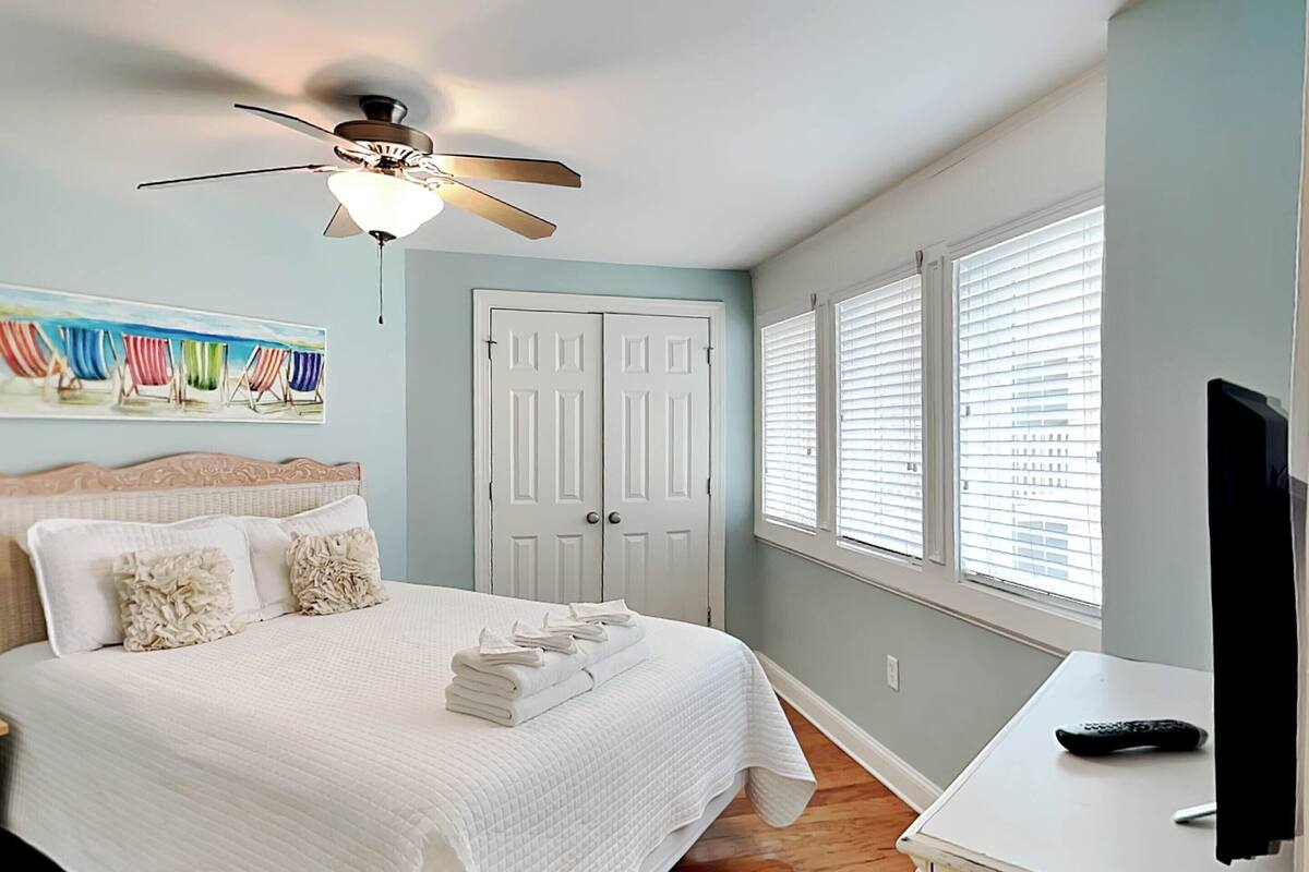 Un dormitorio con cama, ventilador de techo, televisor y un cuadro con sillas de playa.