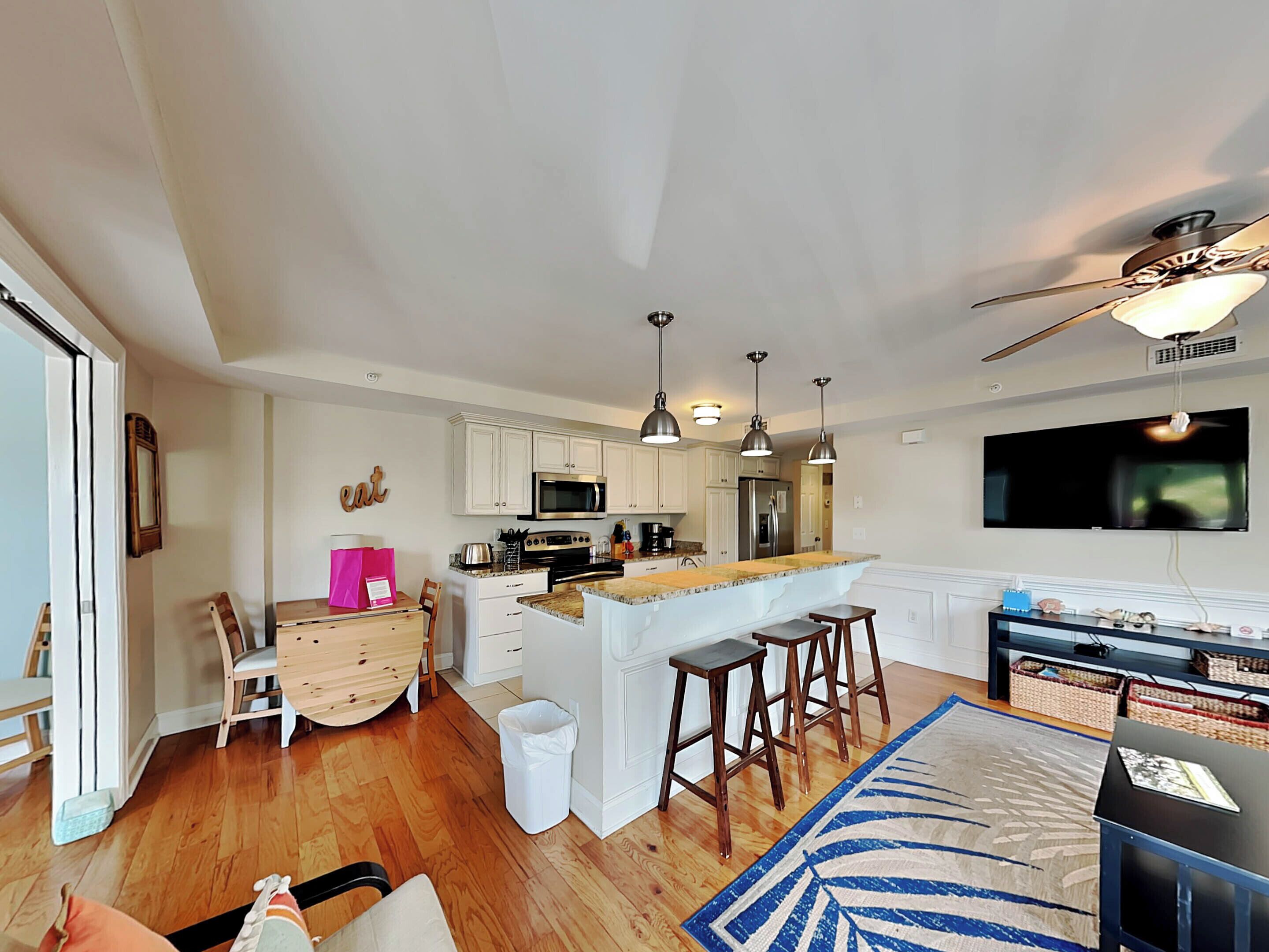 A modern kitchen with white cabinets, a central island, and pendant lighting. There is a dining area with a wooden table and chairs, and a living space with a sofa and a TV mounted on the wall.