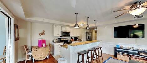 A modern kitchen with white cabinets, a central island, and pendant lighting. There is a dining area with a wooden table and chairs, and a living space with a sofa and a TV mounted on the wall.