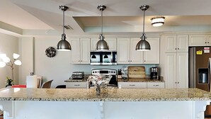 A kitchen with white cabinets, a granite countertop, a stainless steel refrigerator, and three pendant lights. - Captains Watch 1 by Tybee Vacation Rentals (Tybee Island)