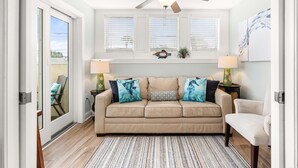 A living room with a beige sofa, two side tables with lamps, a ceiling fan, and a window with blinds. - Captains Watch 1 by Tybee Vacation Rentals (Tybee Island)