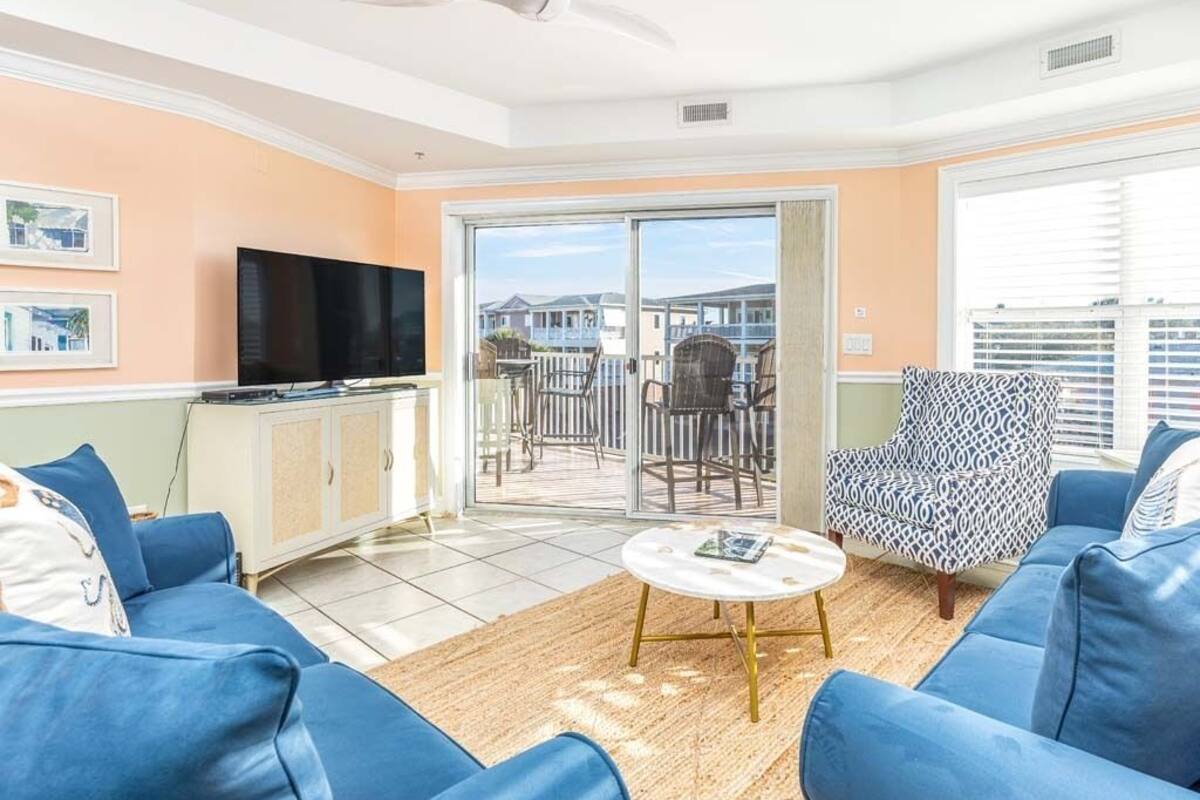 A living room with a TV, blue sofas, a patterned armchair, and a large sliding glass door leading to an outdoor patio.