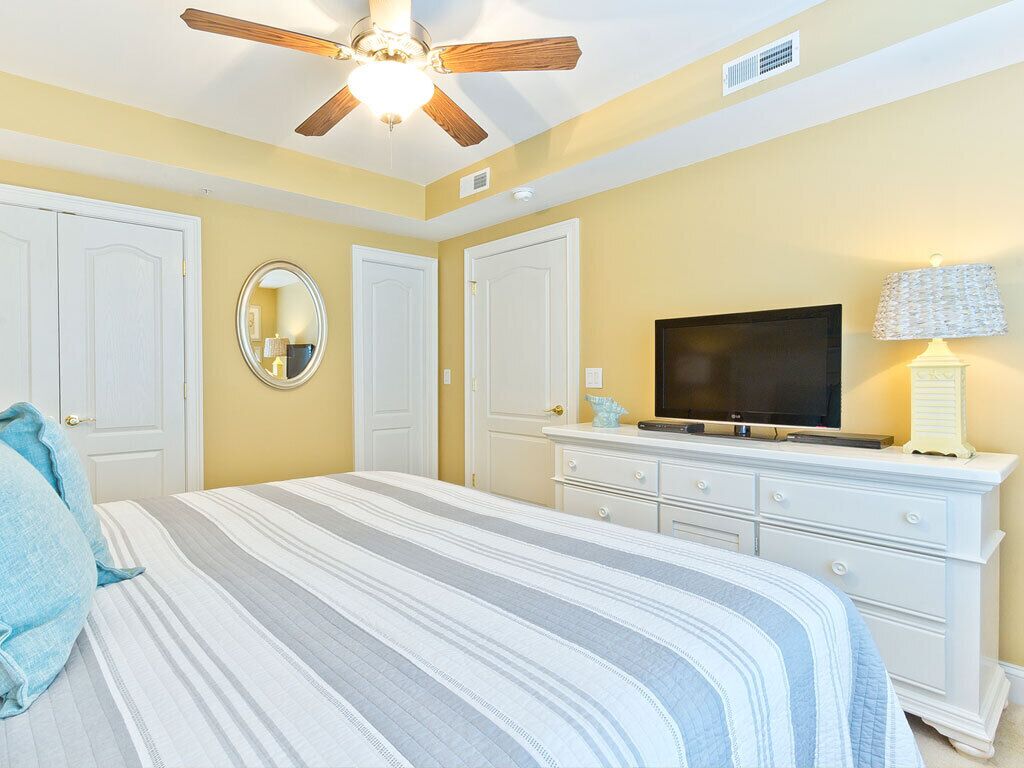 Une chambre à coucher avec un lit, un ventilateur de plafond, une télévision, une commode et un miroir.