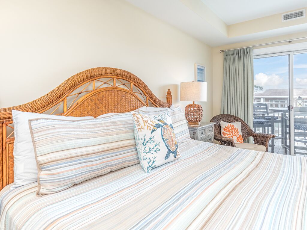 A hotel room with a large bed, a wicker headboard, a view of the city through a balcony, and a lamp on the nightstand.