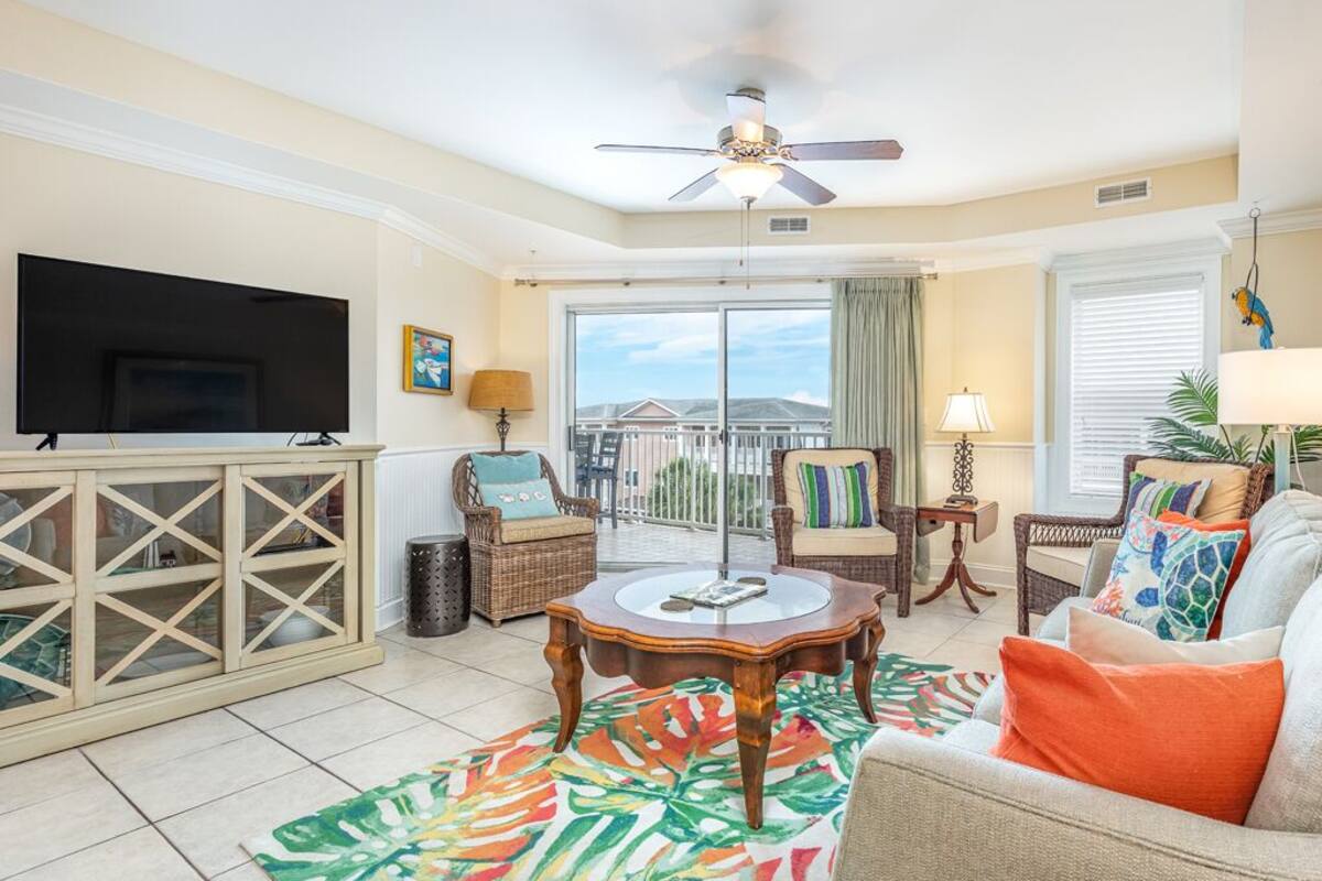A living room with a flat-screen TV, a ceiling fan, wicker furniture, a colorful rug, and a view of the outdoors.