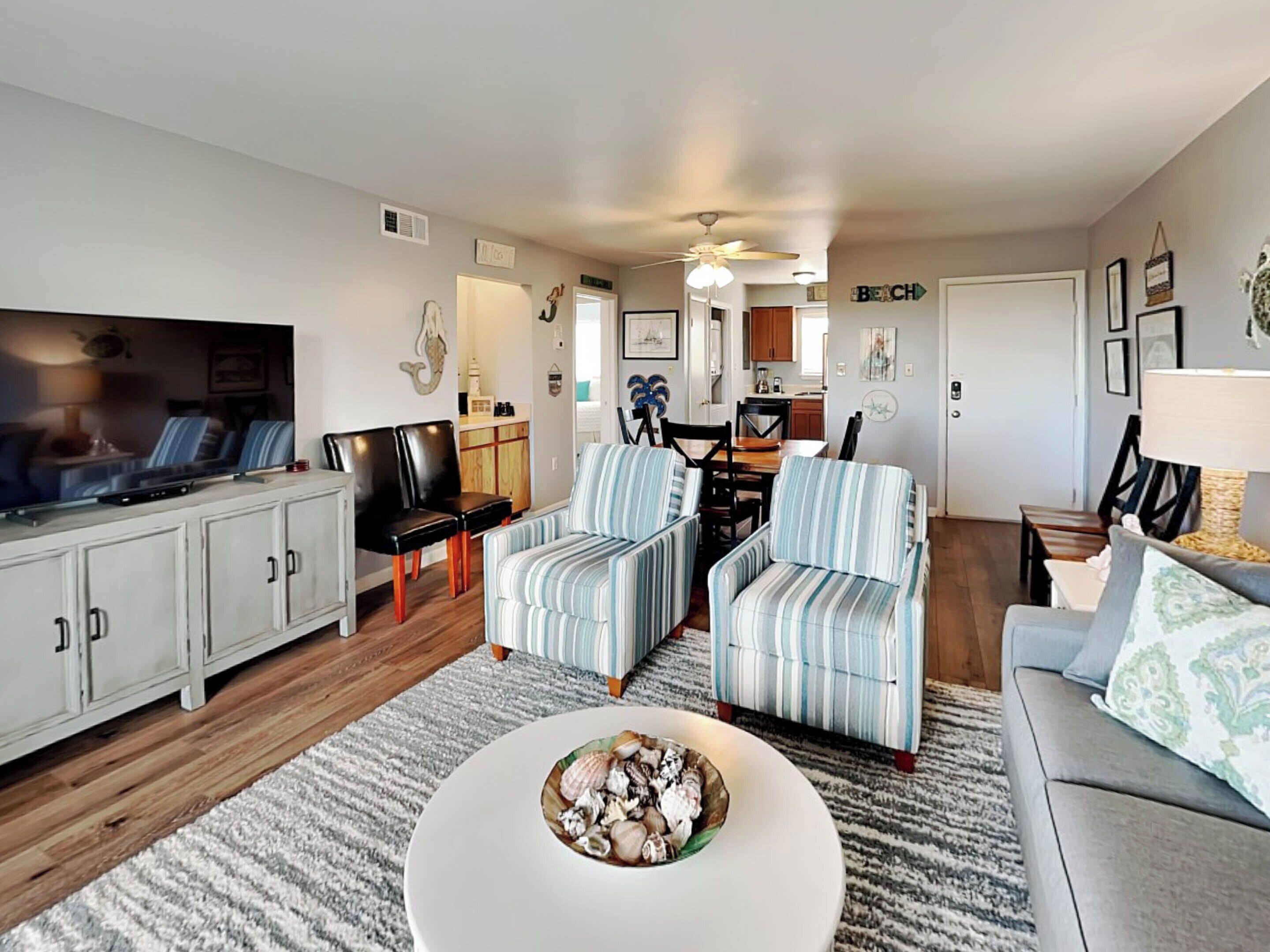 A living room with a television, sofa, striped chairs, a coffee table with seashells, and a kitchen in the background.