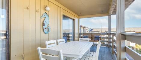 Ein überdachter Außenbereich mit weißem Tisch und Stühlen, einer Seepferdchen-Dekoration und Blick auf den Strand.