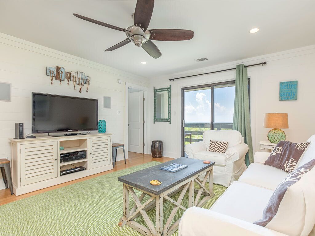 A living room with a television, a ceiling fan, a sofa, a coffee table, and a view of the outdoors.