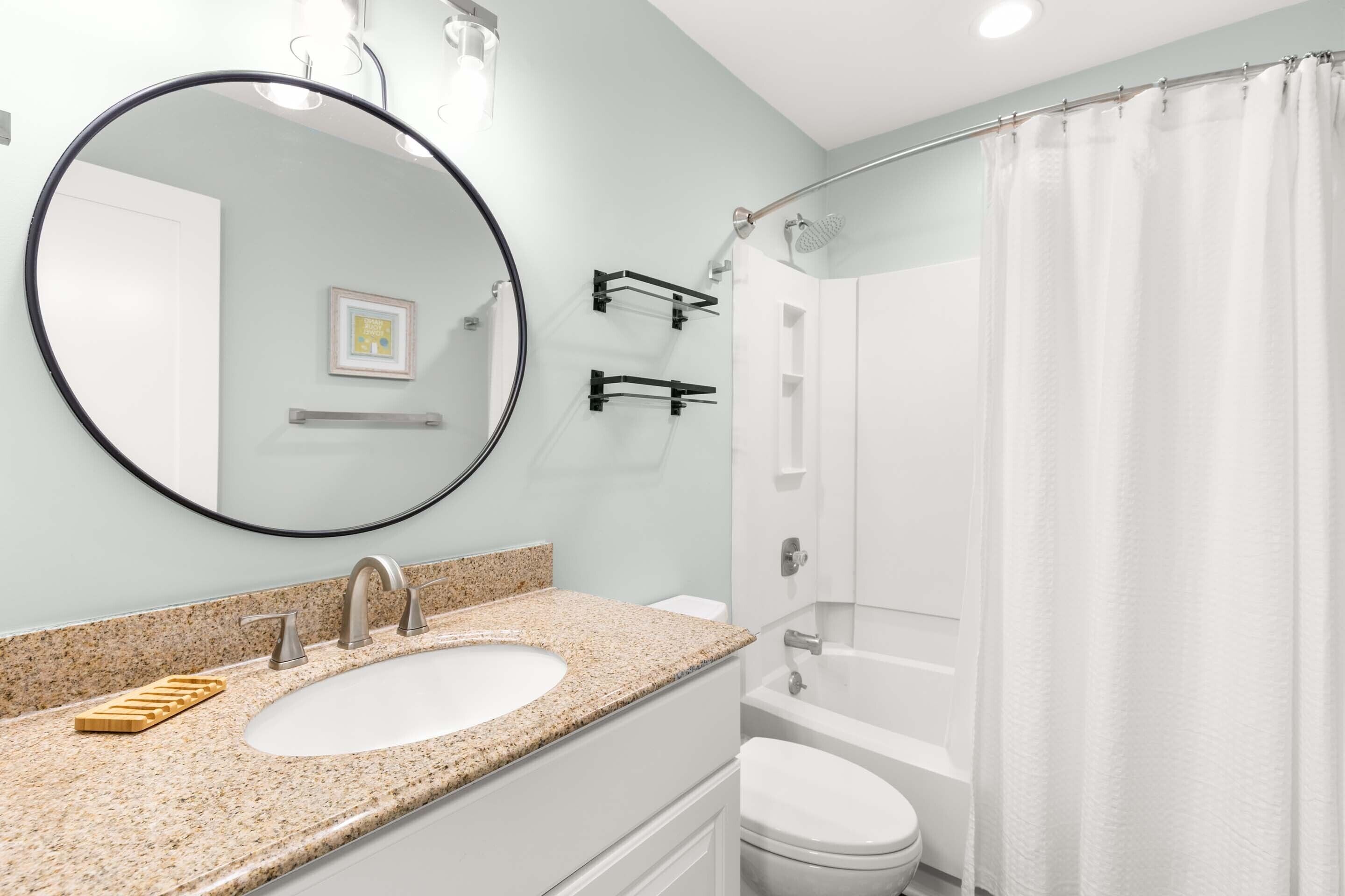 A bathroom with a granite countertop sink, a round mirror, a white toilet, and a glass shower enclosure.