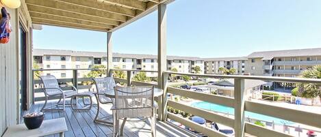 A wooden deck with outdoor seating and a view of a swimming pool and buildings in the background.