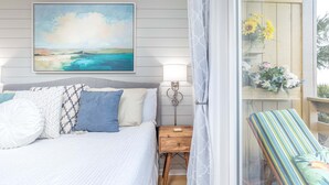 A bedroom with a bed, pillows, a bedside table, a nightstand, a wall-mounted lamp, and a window with curtains. - Lighthouse Point 1 by Tybee Vacation Rentals (Tybee Island)