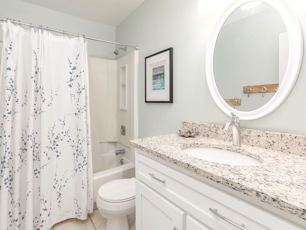Une salle de bain avec des toilettes blanches, un rideau de douche à motifs floraux, un comptoir en granit, un lavabo avec un miroir et un tableau encadré au mur.