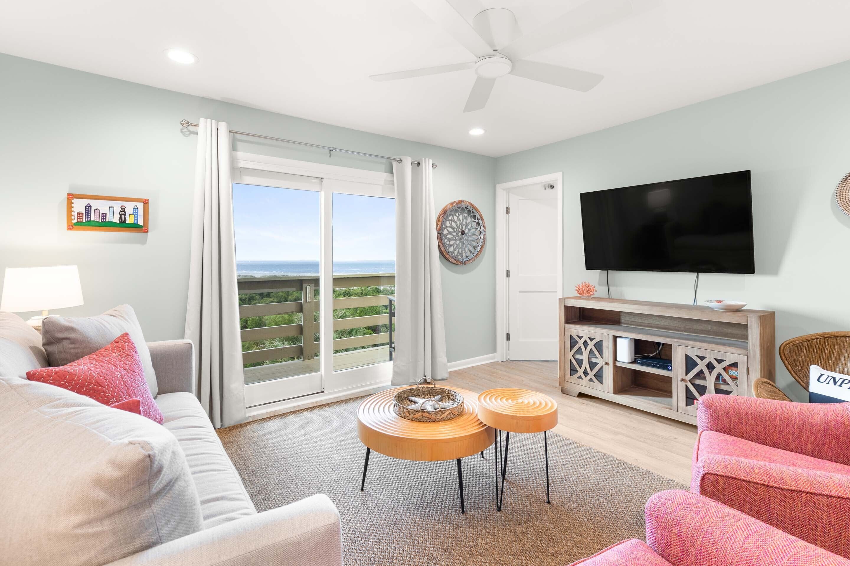 A living room with a sofa, two round wooden side tables, a television, and a balcony with a view.