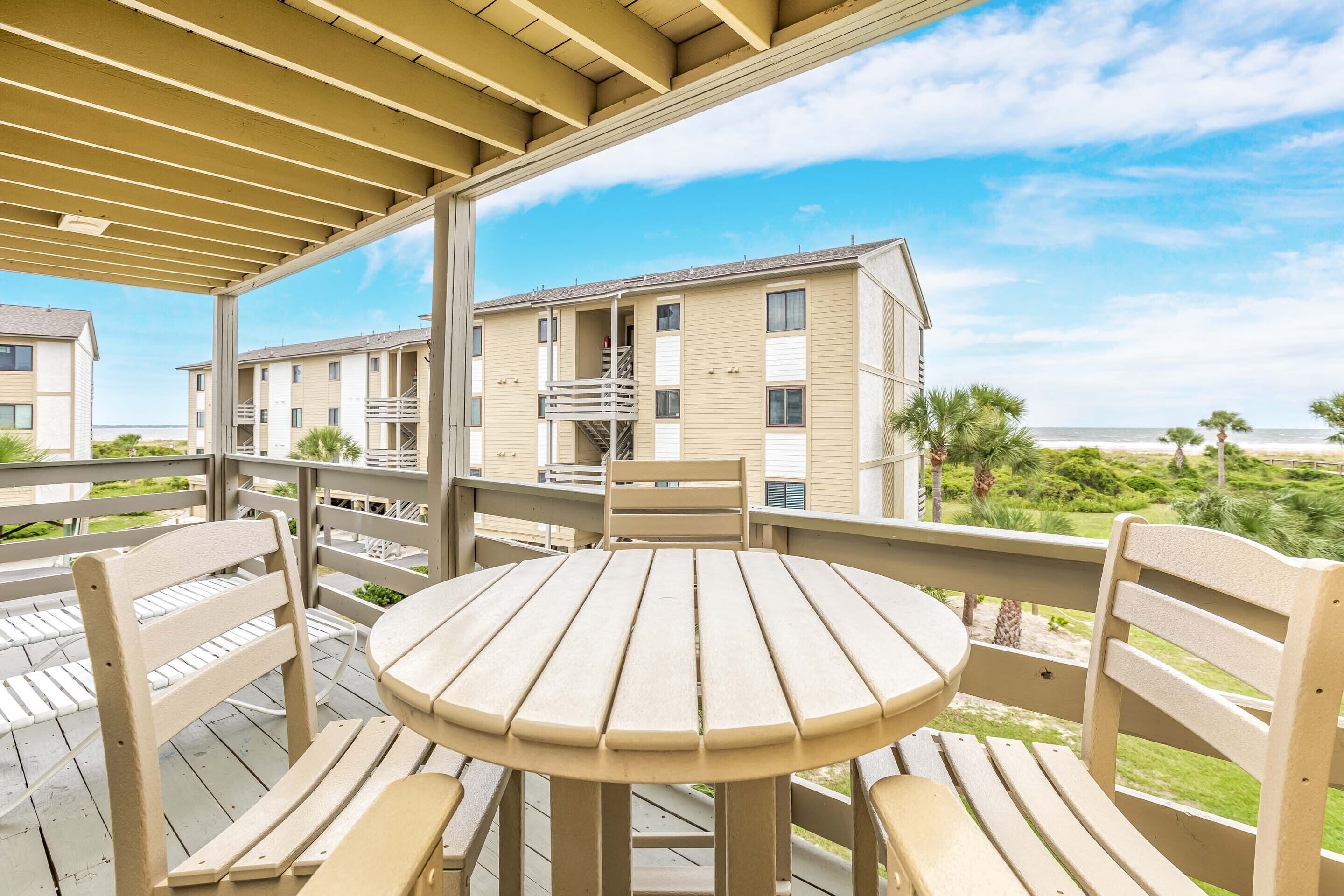 A wooden deck with a round table and chairs, overlooking a building with multiple balconies and a view of the ocean.
