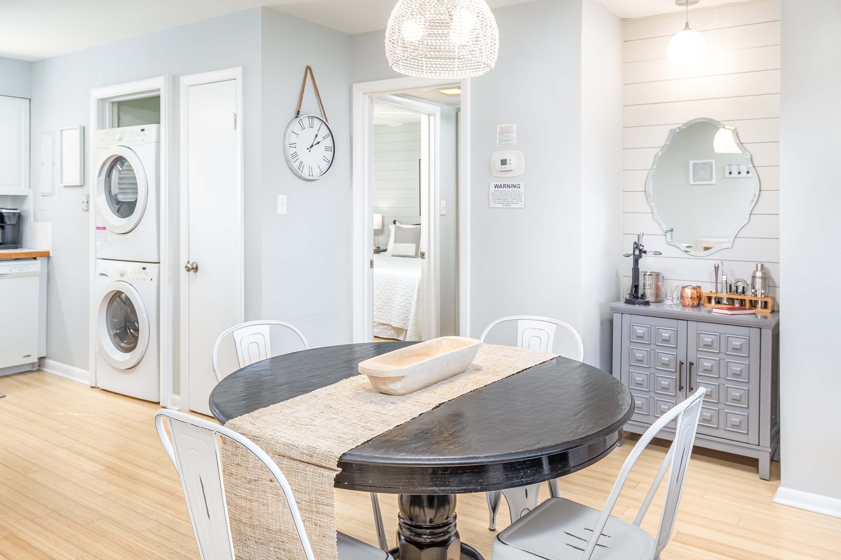 A modern kitchen with a built-in washer and dryer, a dining area with a round table and chairs, and a gray cabinet with a mirror above it.