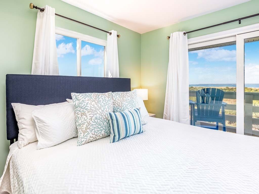Une chambre avec un grand lit, du linge de lit blanc et une vue sur la plage par la fenêtre.
