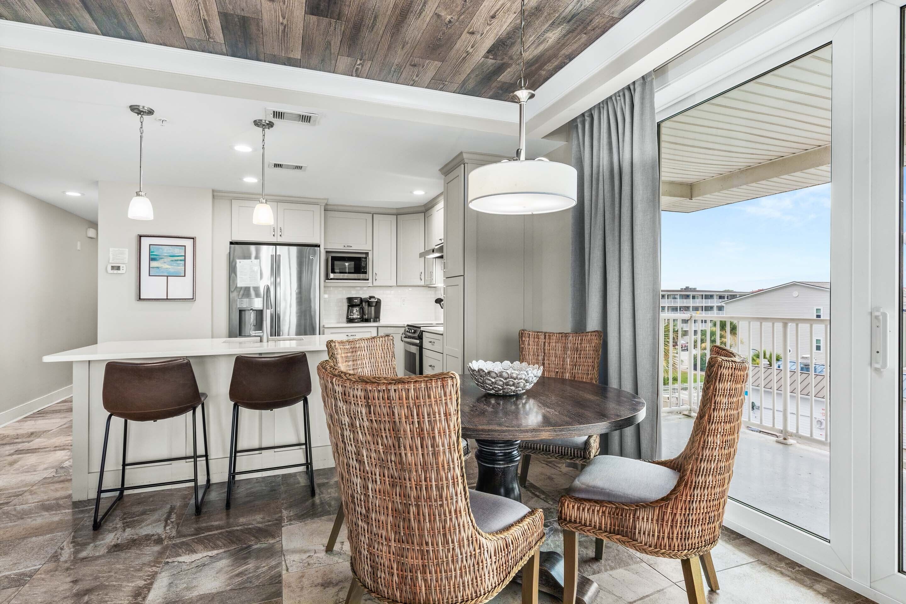 A modern kitchen with stainless steel appliances, a dining area with wicker chairs, and a large window overlooking a balcony.