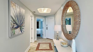 A hallway with a tiled floor, a patterned rug, a white console table with a mirror and a lamp, and a large abstract painting on the wall. - Ocean Song by Tybee Vacation Rentals (Tybee Island)