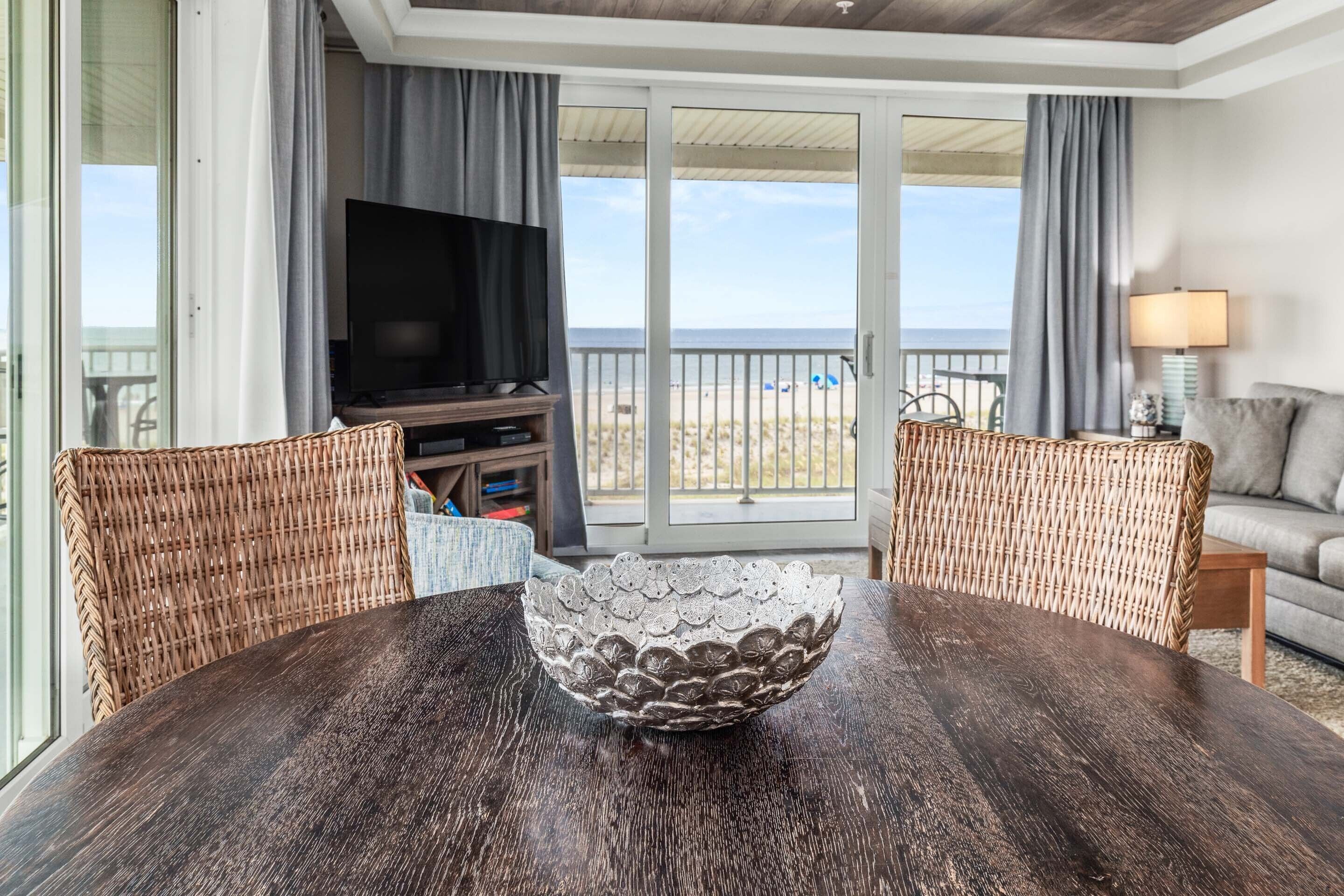 A dining area with a dark wooden table, wicker chairs, a decorative bowl, a television, and a view of the beach through large sliding glass doors.