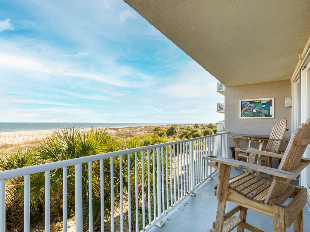 A balcony with a view of the beach and ocean, wooden chairs, and a painting on the wall.
