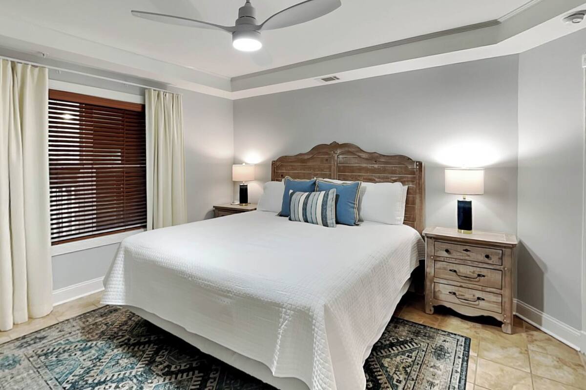 Un dormitorio con una cama grande, cabecera de madera, dos mesitas de noche con lámparas, un ventilador de techo y una ventana con persianas.