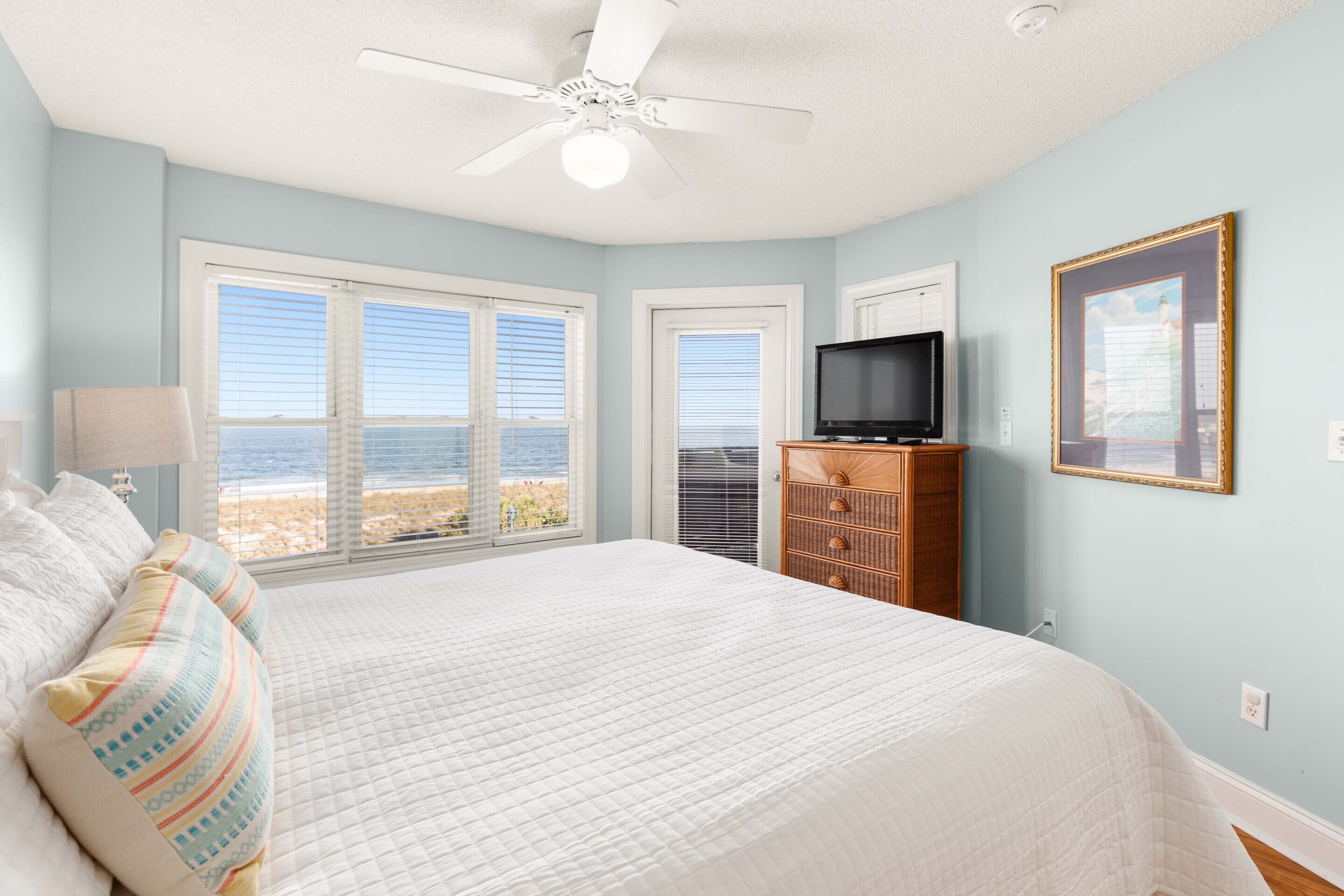 Quarto com cama grande, ventilador de teto, televisão e vista para a praia.