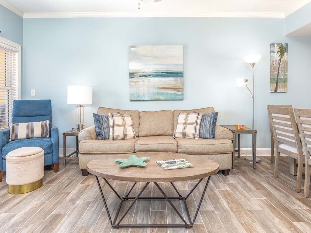 A living room with a beige sectional sofa, a round coffee table, a blue armchair, and a wooden dining table.