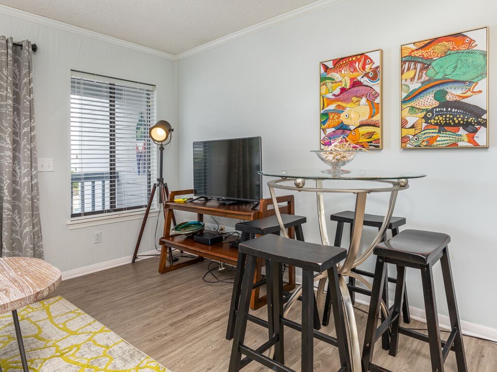 A living room with a TV, a glass table, two bar stools, a sofa, and a window with blinds.
