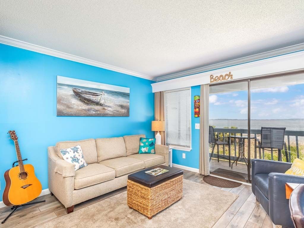 A living room with a blue wall, a beige sofa, a wicker coffee table, and a guitar. There's a balcony with a view of the ocean.