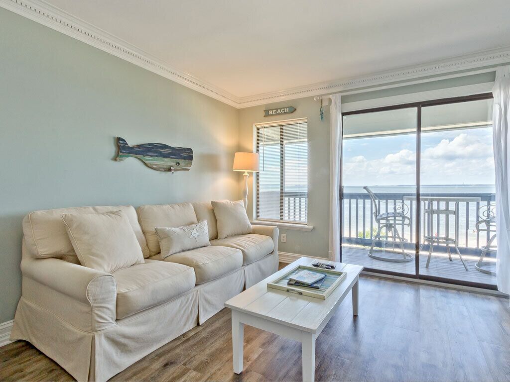 A living room with a beige sofa, a white coffee table, and a view of the ocean through a sliding glass door.